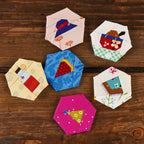 Six colourful quilt blocks arranged on a wooden surface. Each hexagonal piece features varied patterns, textures, and bright hues, creating a playful and crafty vibe.