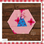 Hexagon-shaped fabric patch on wood, pink with floral patterns and a blue triangular section with red dots, adorned with a pink tassel. Creative, crafty tone.
