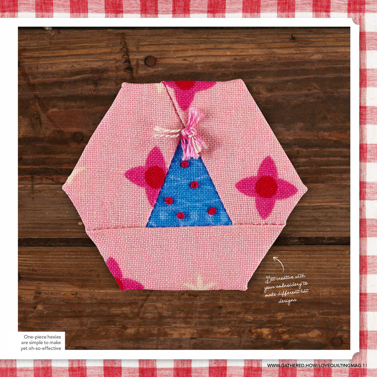 Hexagon-shaped fabric patch on wood, pink with floral patterns and a blue triangular section with red dots, adorned with a pink tassel. Creative, crafty tone.