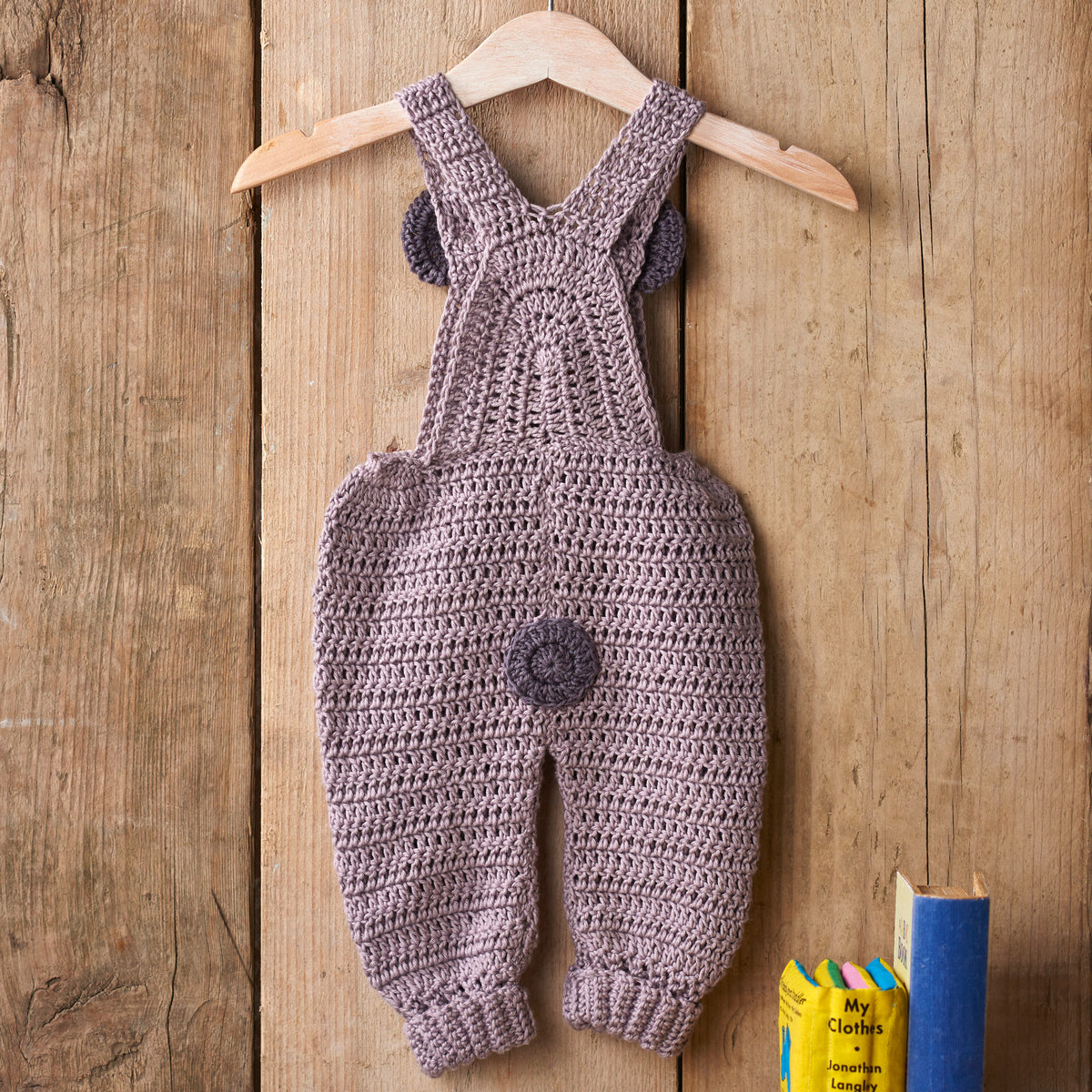 Crocheted baby overalls on a wooden hanger against a wooden background