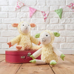 Two fluffy toy sheep with a red suitcase against a white brick wall with colorful bunting.