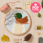 Embroidery hoop with a completed embroidery design of a person wearing a green hat and brown scarf, surrounded by sewing supplies on a beige background.