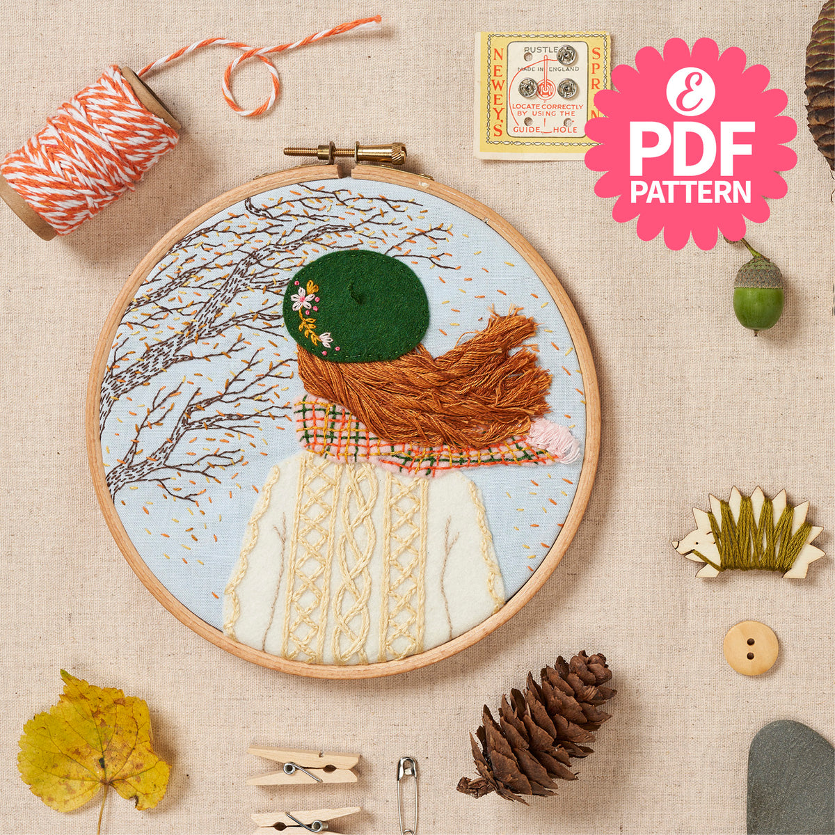 Embroidery hoop with a completed embroidery design of a person wearing a green hat and brown scarf, surrounded by sewing supplies on a beige background.
