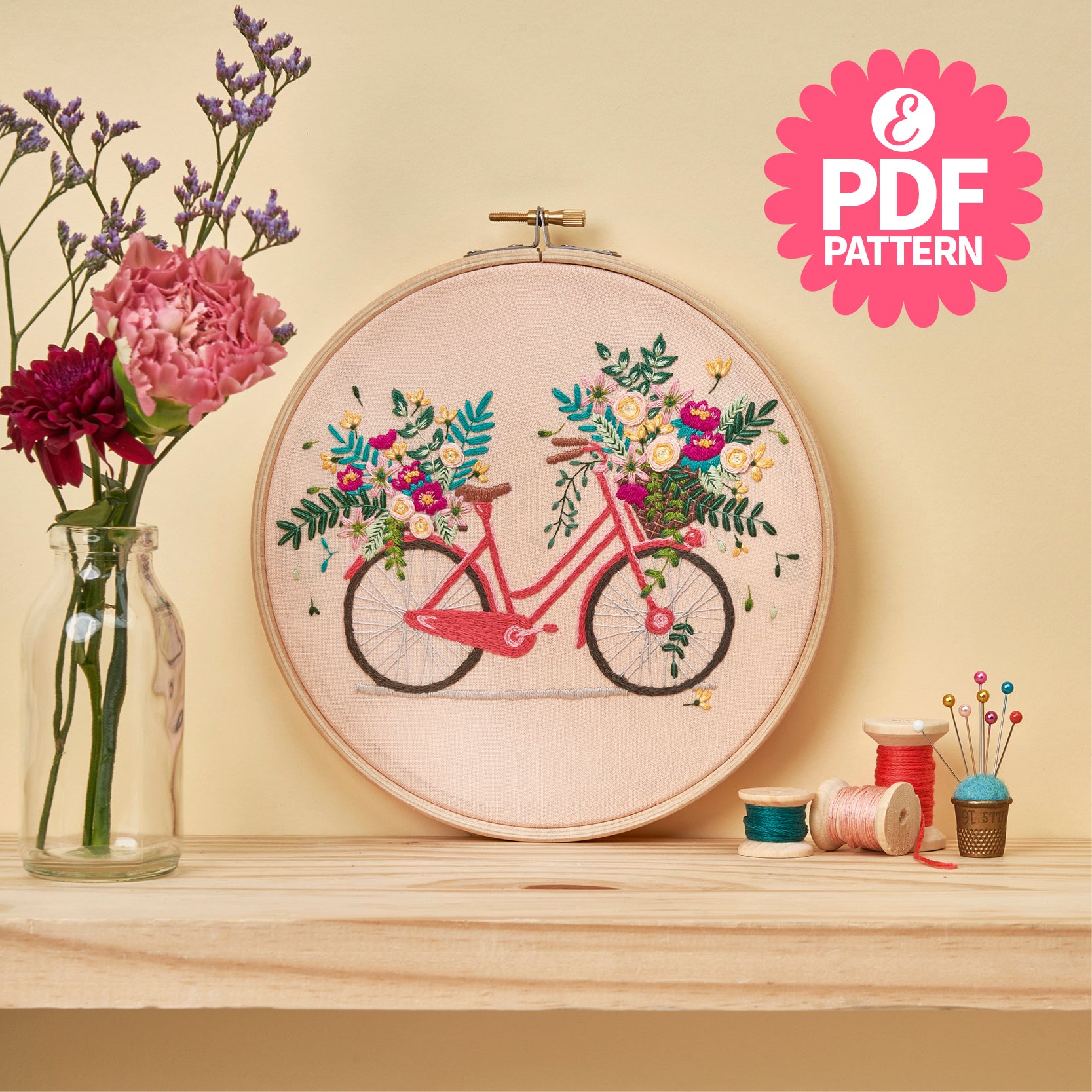 Embroidery hoop with bicycle and floral design on a wooden surface with flowers and thread spools.