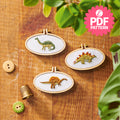 Three embroidered dinosaur designs in wooden hoops on a wooden surface with a PDF pattern badge.