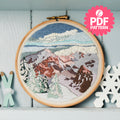 Embroidery hoop with mountain landscape design on a white wooden shelf.