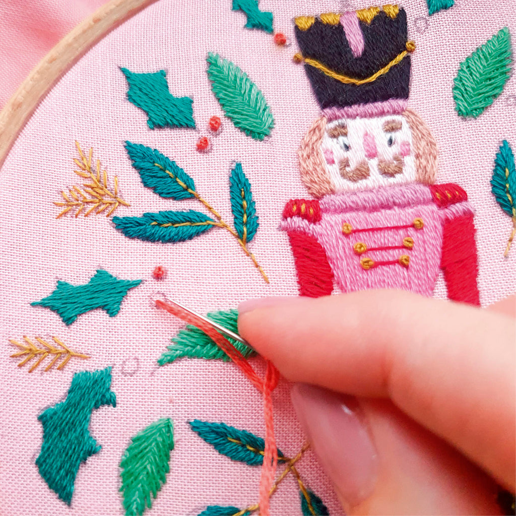 Embroidery hoop with a nutcracker design and green leaves, with a hand holding a needle.