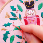 Embroidery hoop with a nutcracker design and green leaves, with a hand holding a needle.