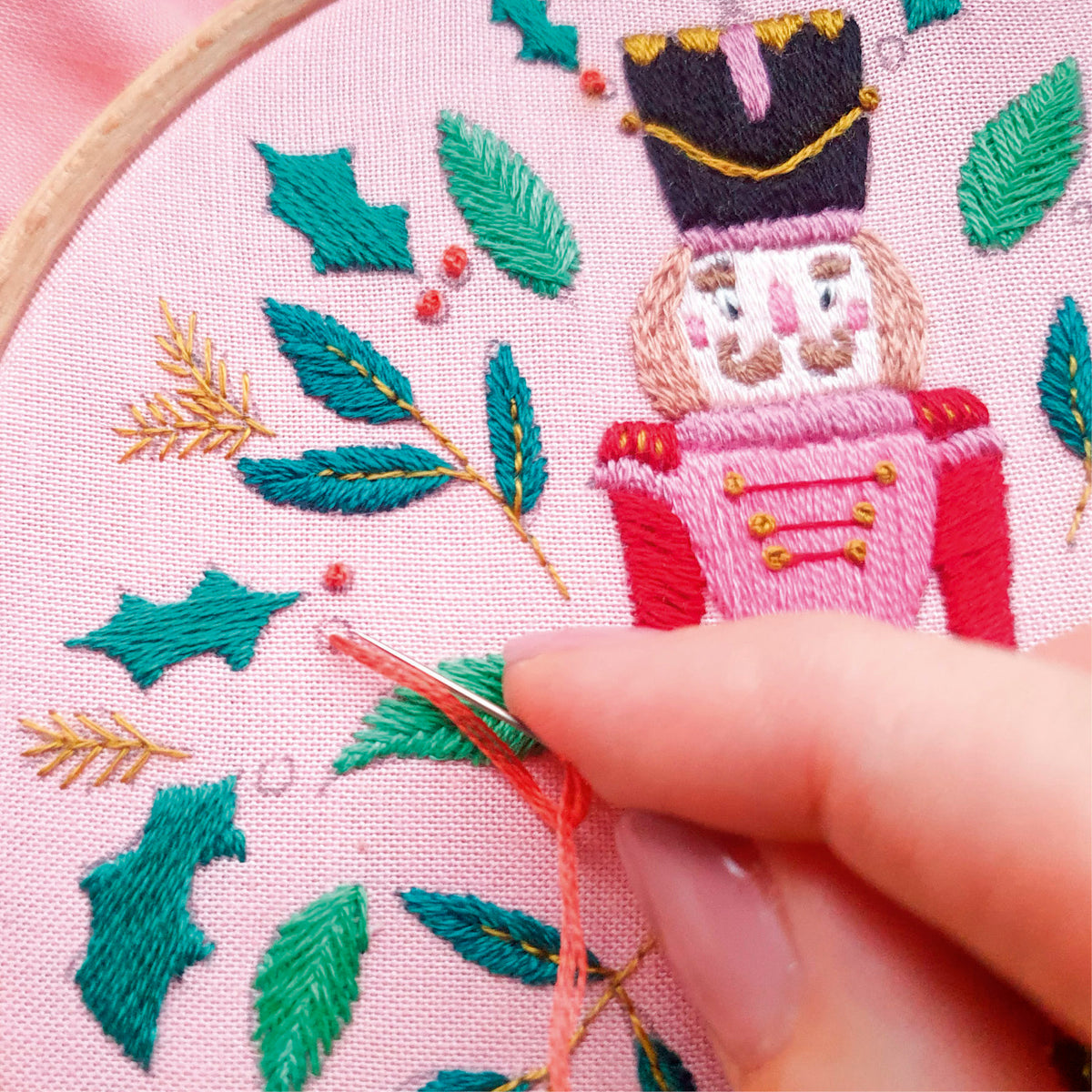 Embroidery hoop with a nutcracker design and green leaves, with a hand holding a needle.