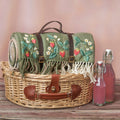 Wicker picnic basket with embroidered green blanket, bottles of pink juice on a wooden surface.