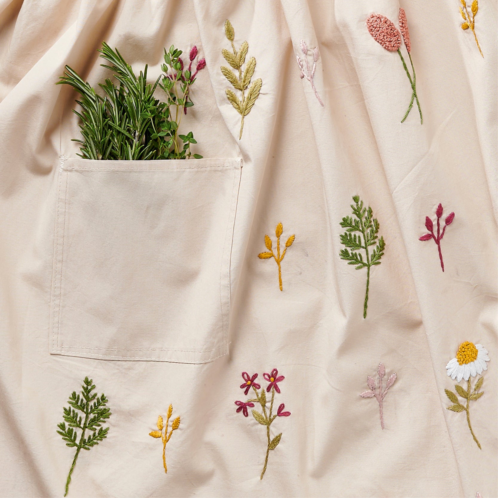 Beige apron with embroidered plants and a pocket containing herbs