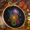 Embroidery hoop with embroidered fireworks design on a dark background, surrounded by autumn leaves and lights.
