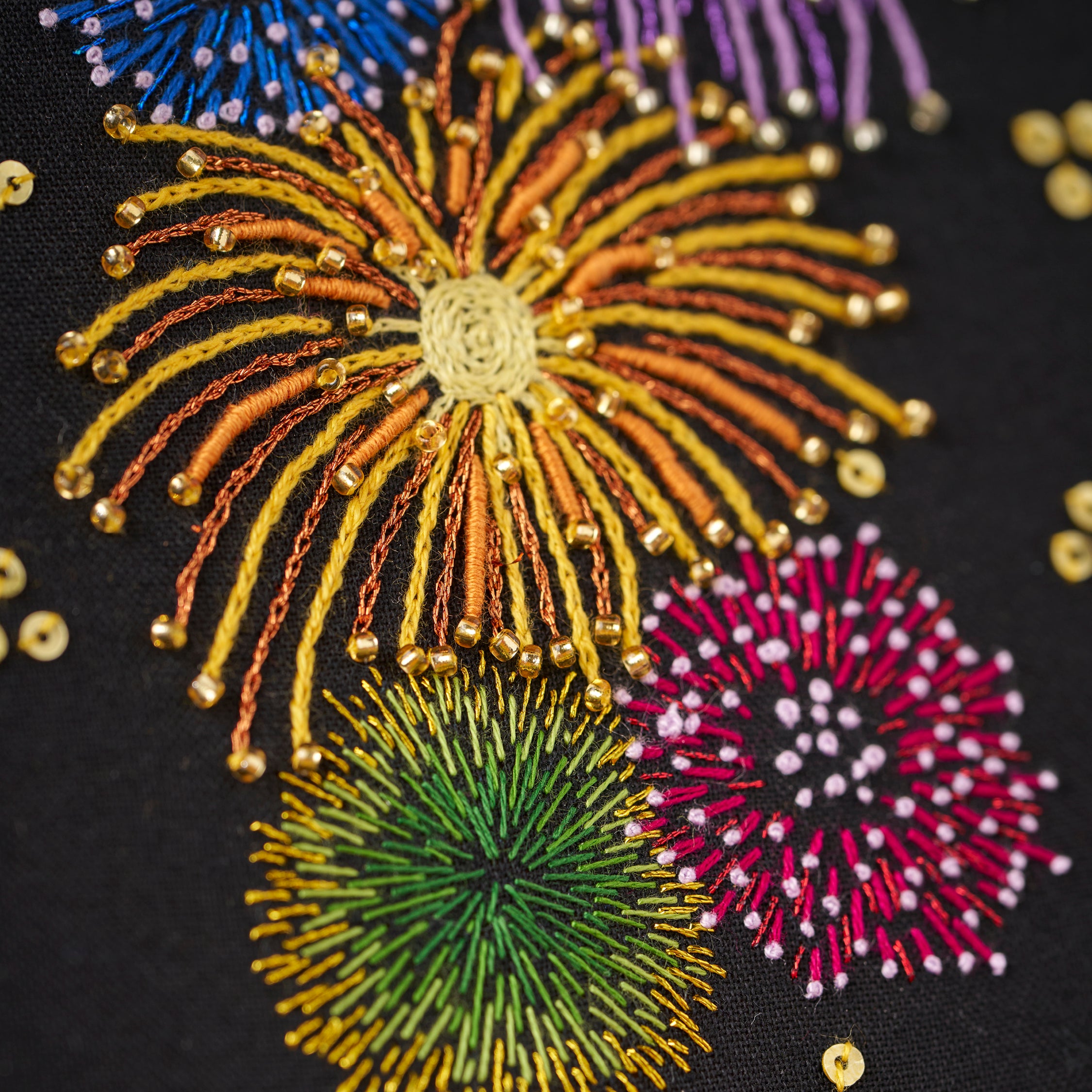 Embroidery hoop with embroidered fireworks design on a dark background, surrounded by autumn leaves and lights.
