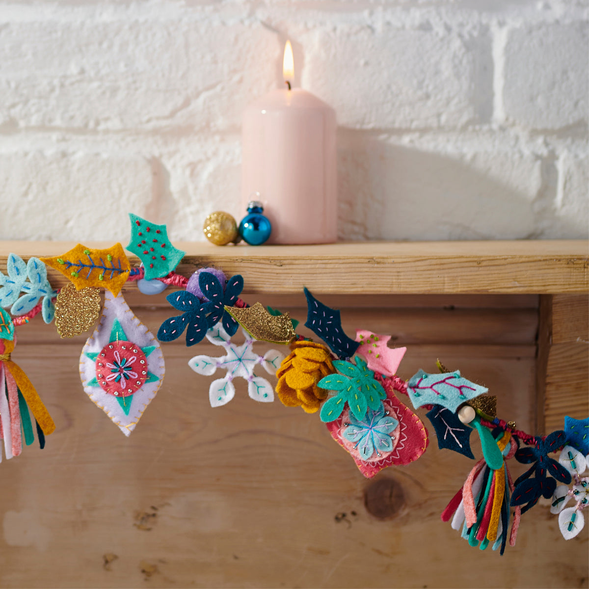 Decorative garland with colorful tassels and flowers on a wooden mantel with a lit pink candle.