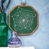 Decorative spiderweb embroidery in a wooden frame on a textured wall with two vases.