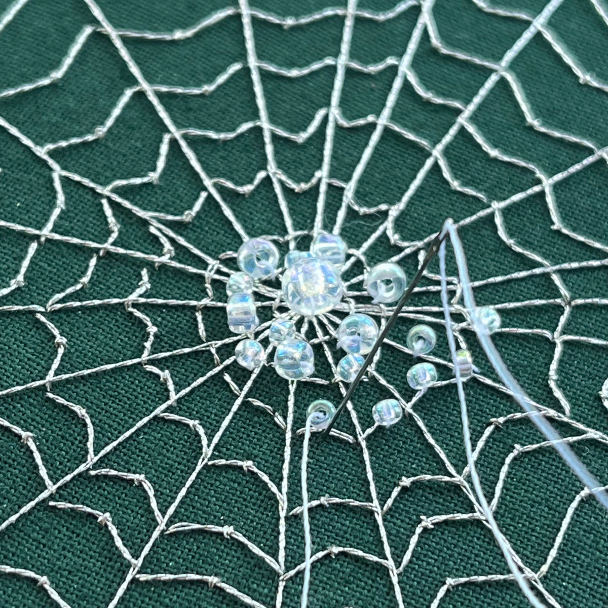 Close-up of a spiderweb embroidery  with a decorative bead in the center on a green fabric background.