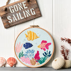 Embroidery hoop with a colorful underwater scene, 'Gone Sailing' sign, and decorative items on a white wooden background.