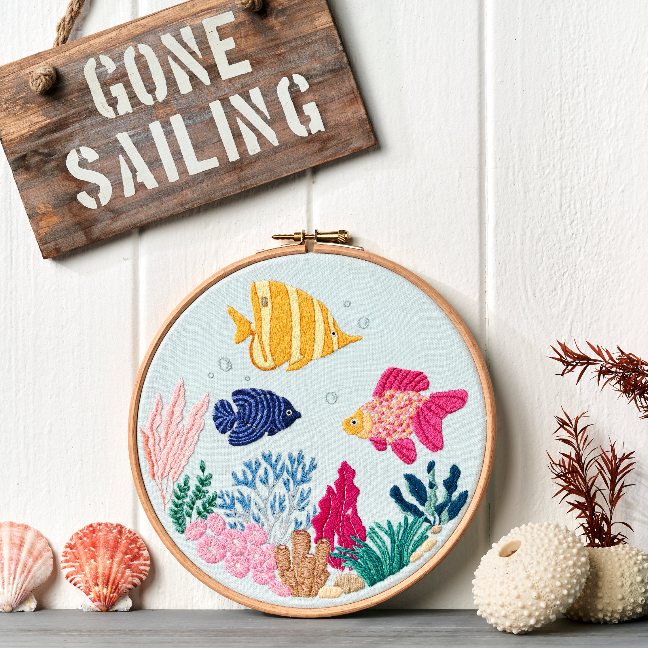Embroidery hoop with a colorful underwater scene, 'Gone Sailing' sign, and decorative items on a white wooden background.