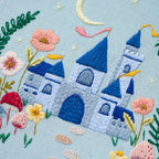 Embroidered design of a castle with flowers and plants on a light blue background