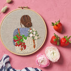 Embroidery hoop with a girl holding a bag of fruits and flowers on a pink background with strawberries and flowers.