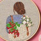 Embroidery hoop with a detailed embroidery of a person wearing a striped shirt with floral and berry designs.