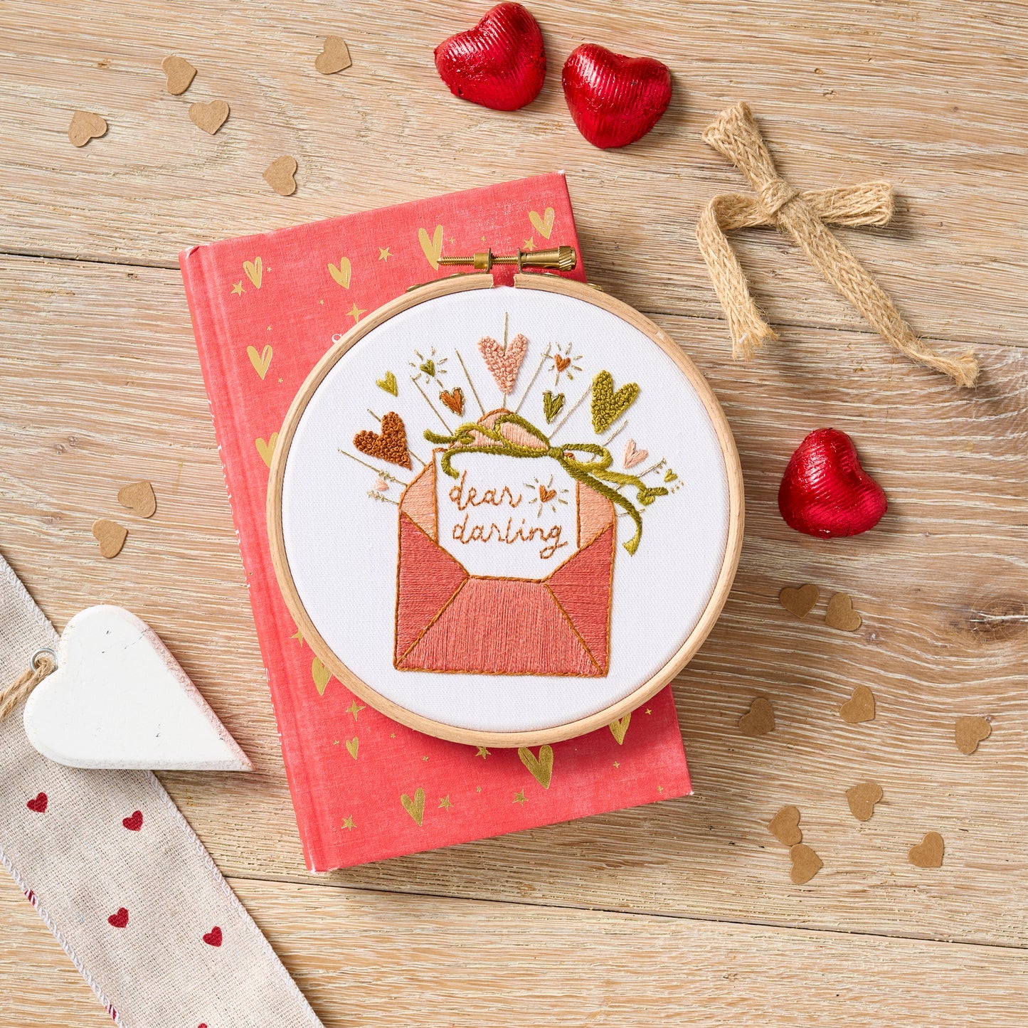 Embroidery hoop with 'dear darling' design on a pink book with heart decorations on a wooden surface.