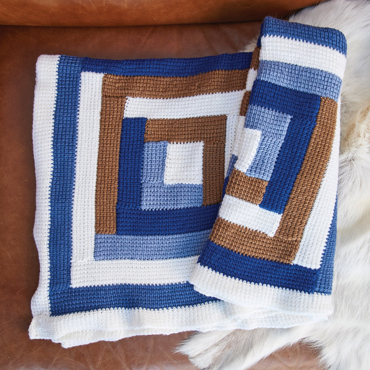 A crocheted blanket with a geometric pattern of blue, brown, and white squares folded on a tan leather couch. The design creates a modern, cozy feel.