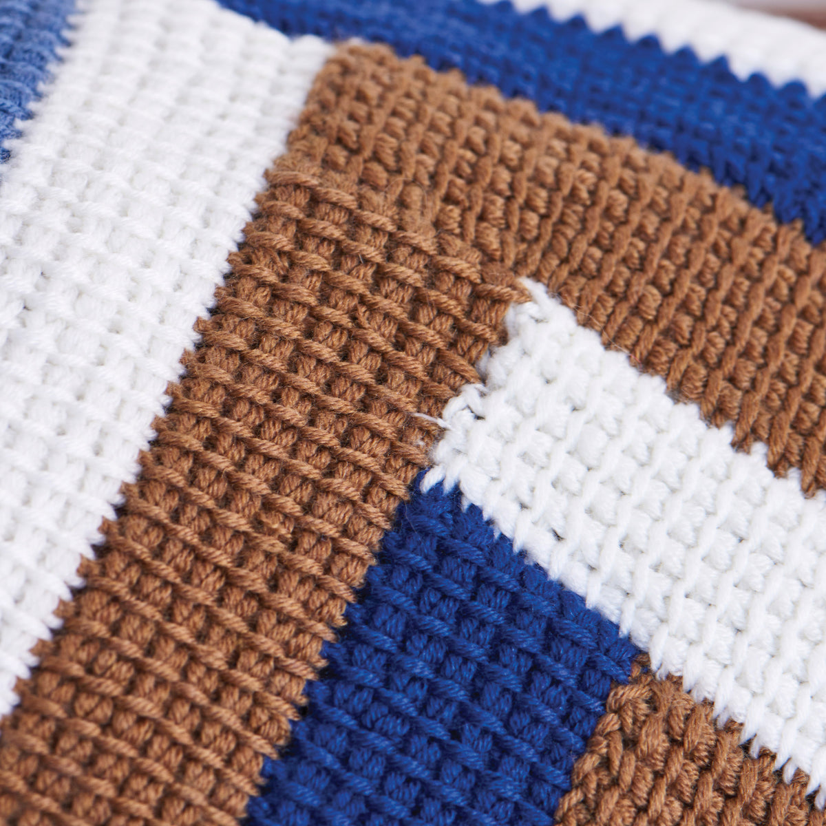 Close-up of a crocheted fabric with a striped pattern in white, brown, and blue. The textured stitching creates a cozy, handcrafted feel.