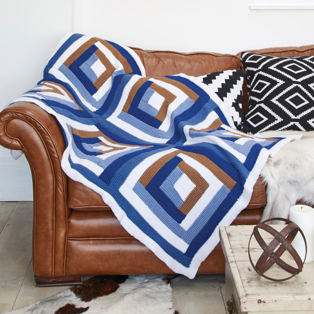A cozy, brown leather sofa is draped with a crocheted blanket featuring blue, white, and brown geometric patterns. Black and white patterned cushions add contrast.