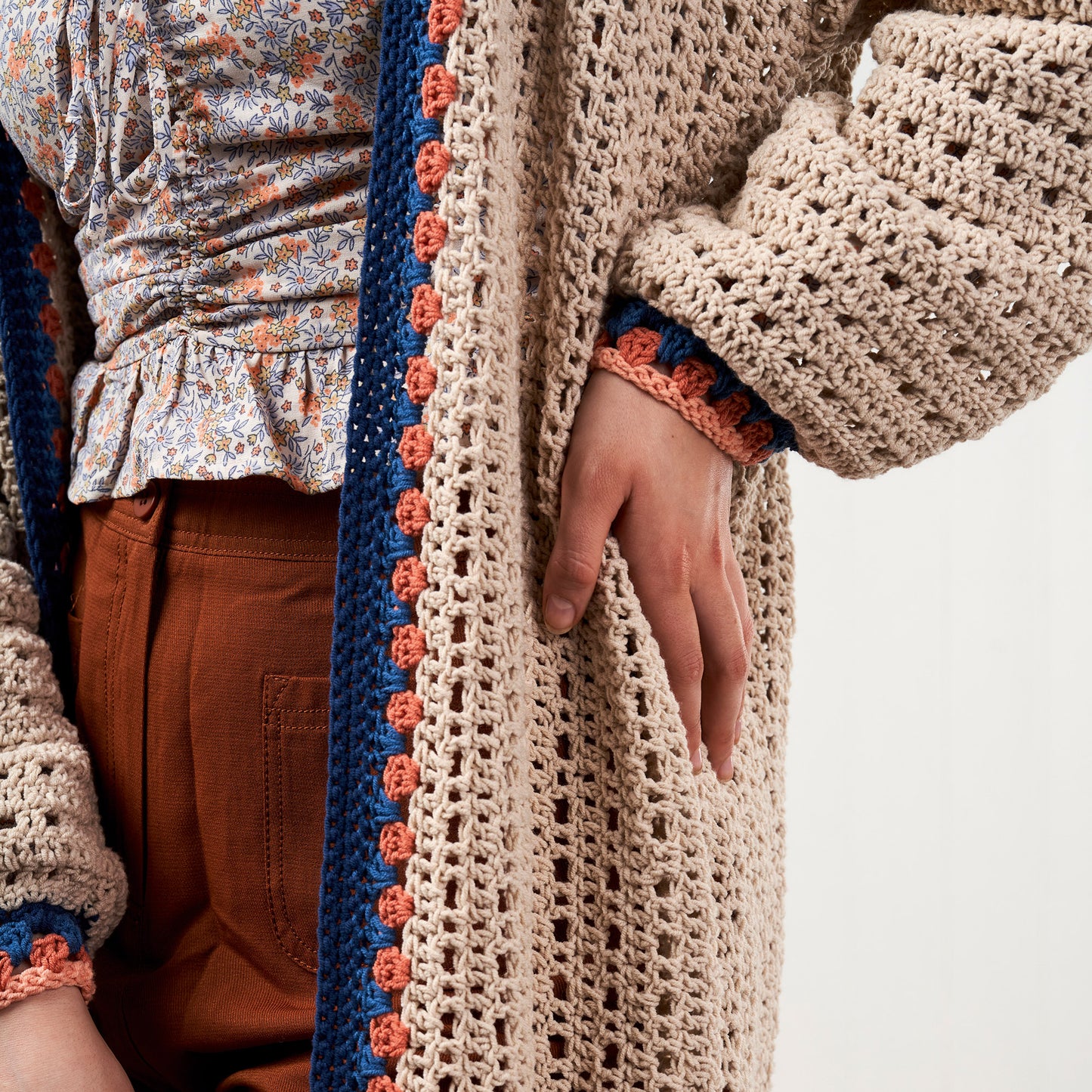 Close-up of a person wearing a beige crochet cardigan with colorful trim on a white background