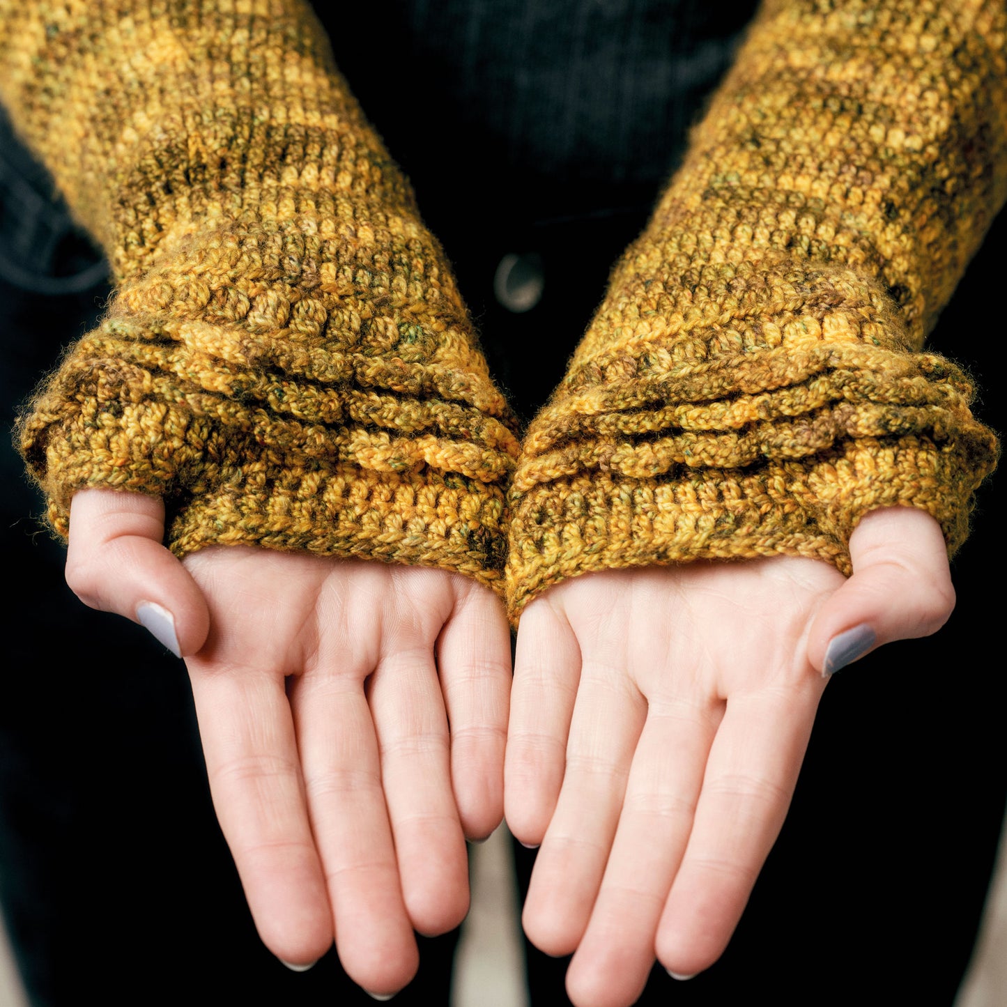 Close-up of hands wearing mustard-yellow knit fingerless gloves. The palms face upward, showcasing the textured stitches. Cozy and warm atmosphere.
