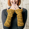 A woman in a gray ribbed sweater smiles warmly, holding her face with hands adorned in mustard-yellow, fingerless crocheted gloves.