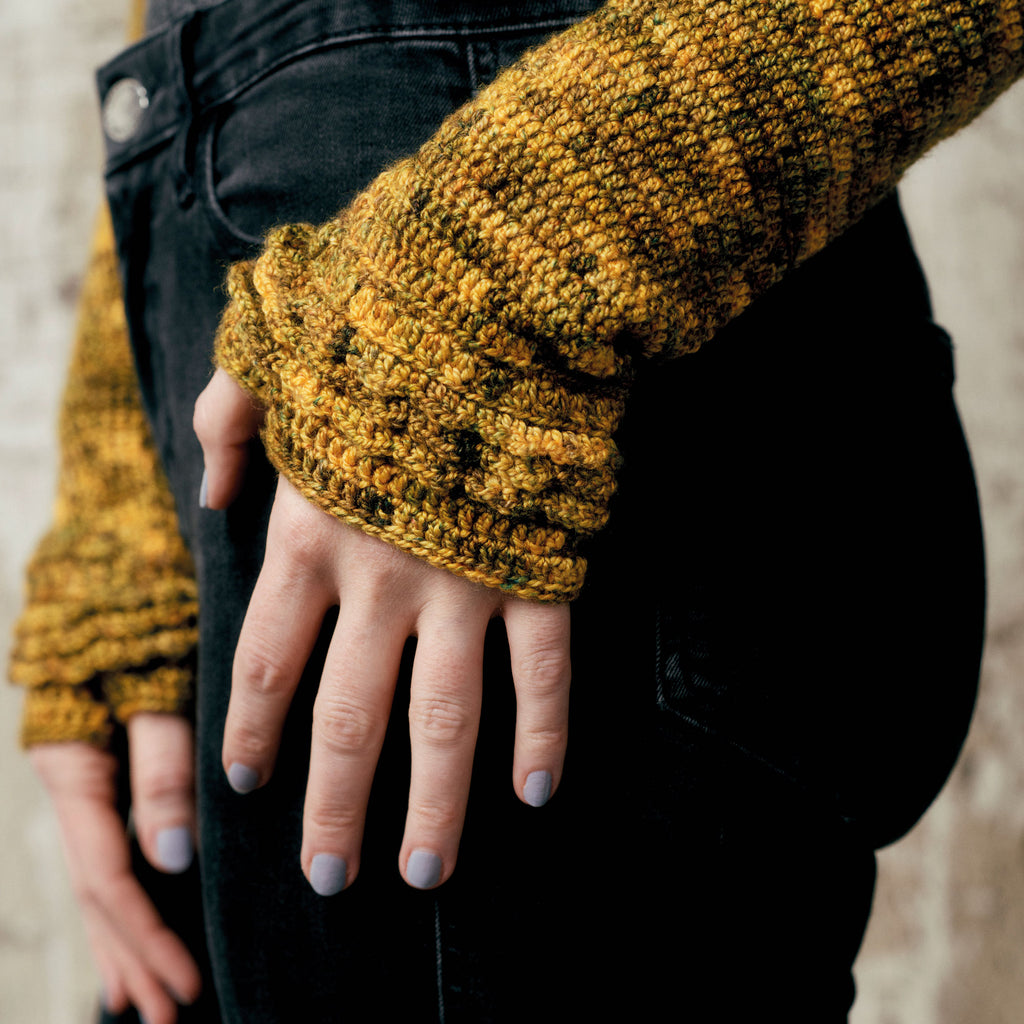 Close-up of a hand against dark jeans, wearing a mustard-yellow crocheted sweater with layered sleeves. The hand displays light purple nail polish.