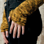 Close-up of a hand against dark jeans, wearing a mustard-yellow crocheted sweater with layered sleeves. The hand displays light purple nail polish.