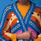 Person wearing a colorful crocheted cardigan with a blue, orange, and yellow pattern against a blue background.