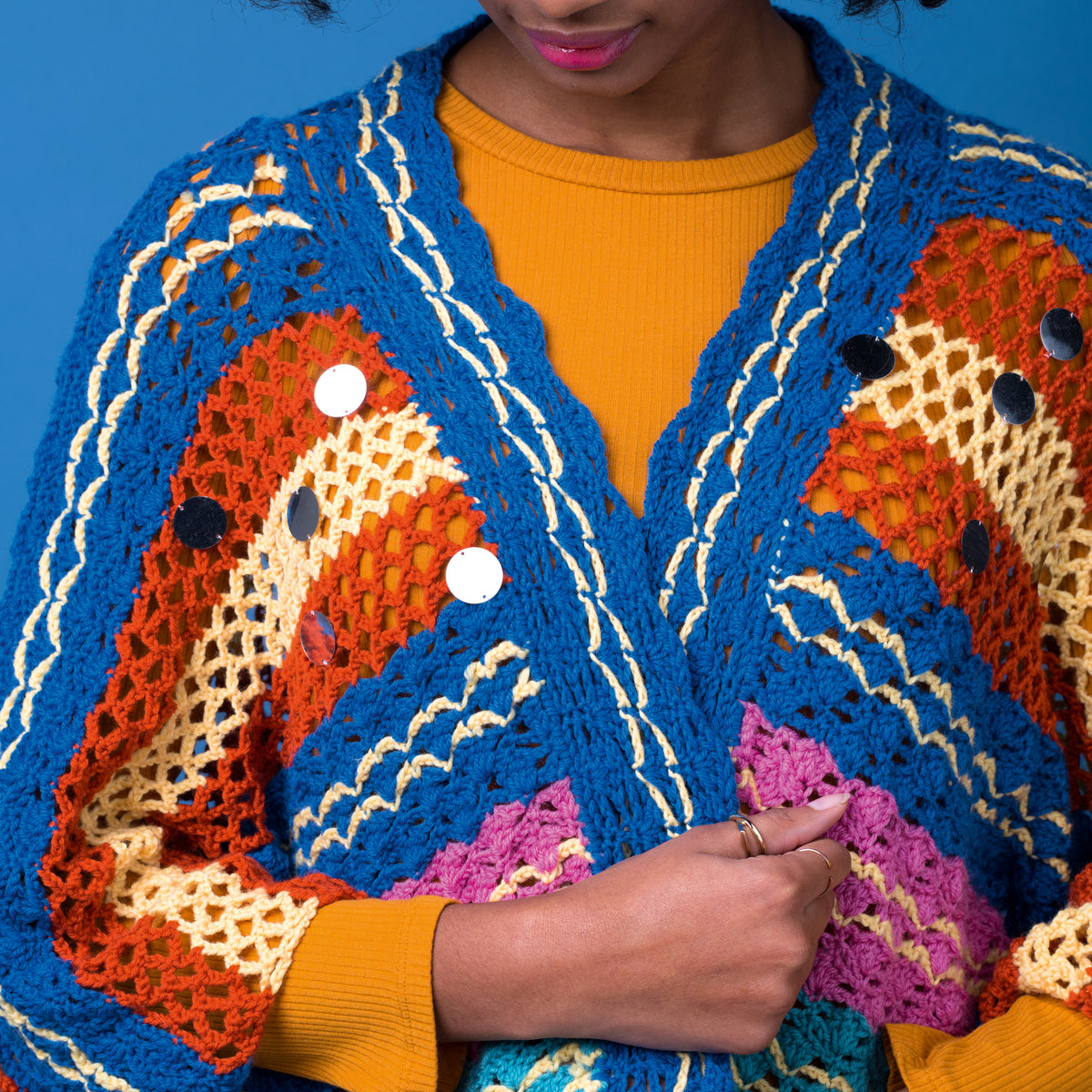 Person wearing a colorful crocheted cardigan with a blue, orange, and yellow pattern against a blue background.