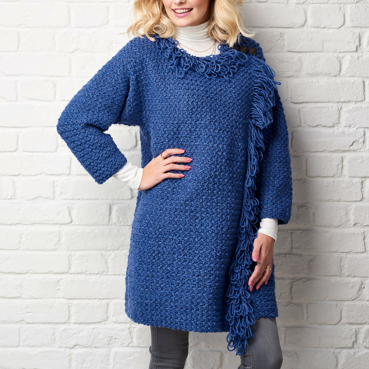 Woman wearing a blue crochet poncho against a white brick wall.