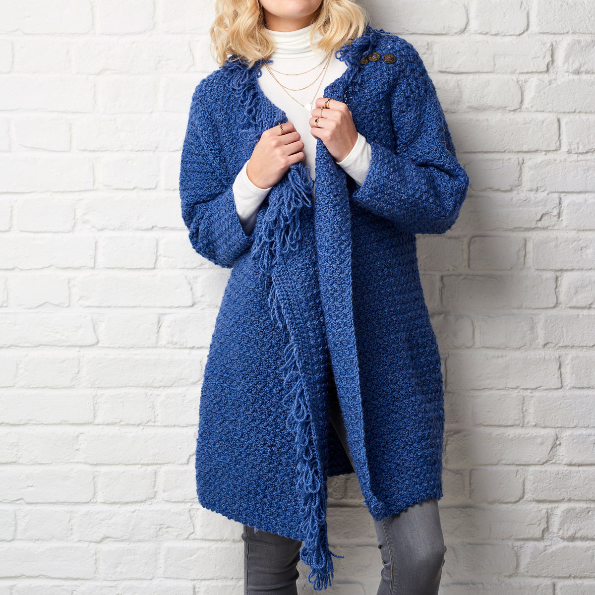 Person wearing a blue crochet cardigan against a white brick wall
