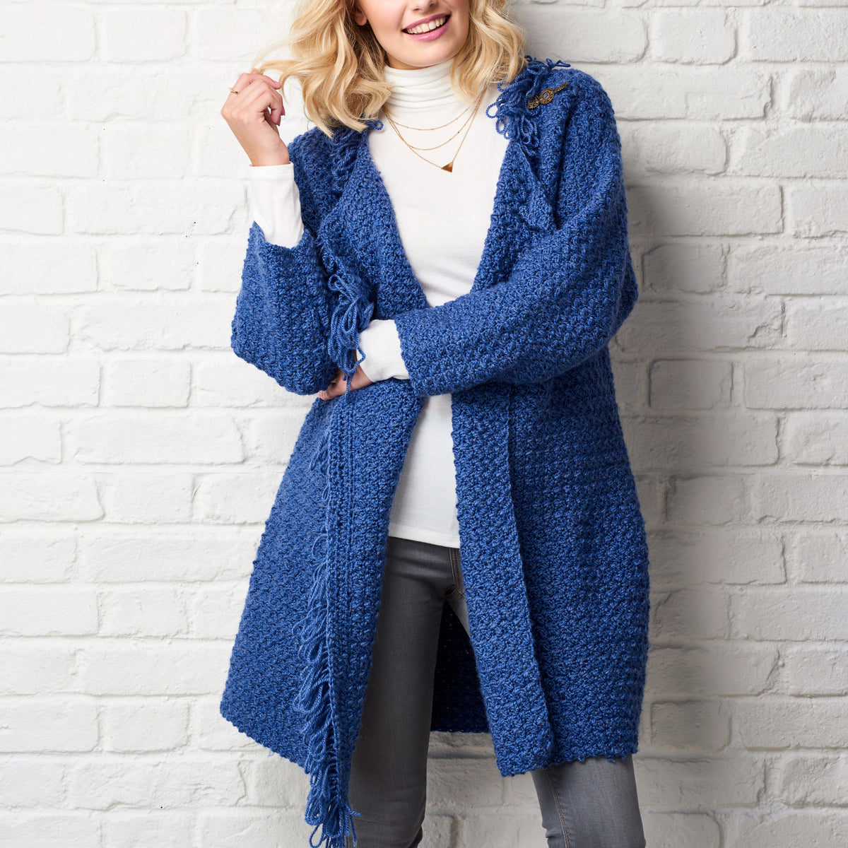 Woman wearing a blue crochet cardigan against a white brick wall
