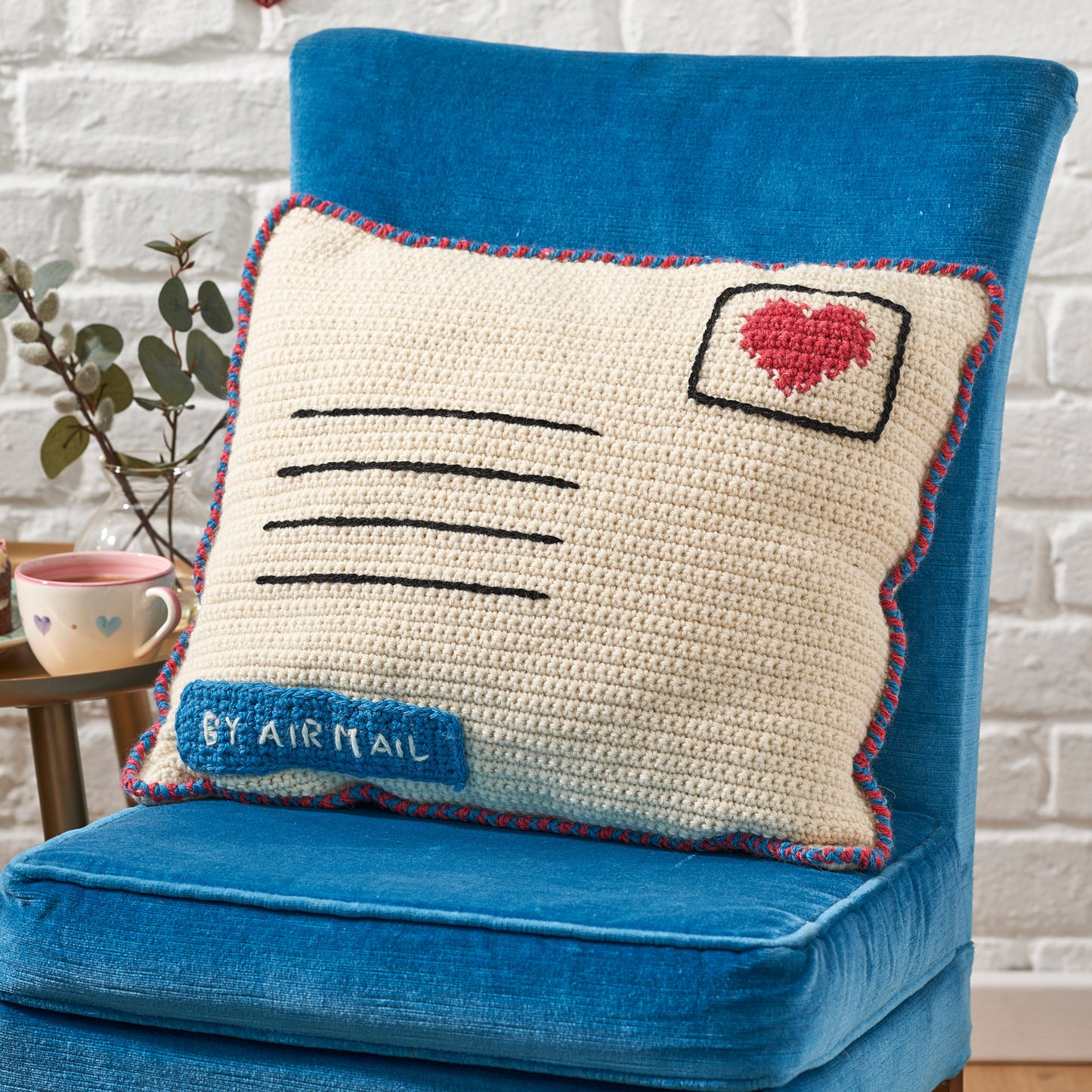 Crocheted letter-shaped pillow with heart and 'By Airmail' text on a blue chair.