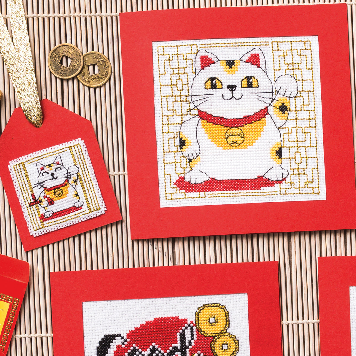 Cross-stitched lucky cat design on red fabric with gold coins and a red tag.