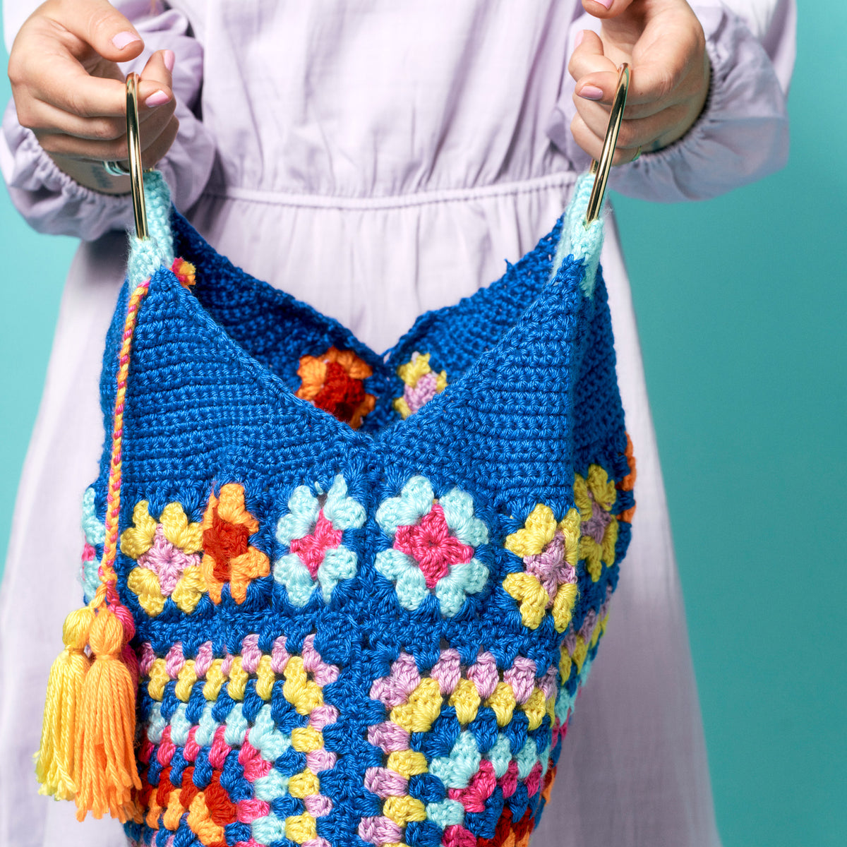 Colorful crocheted bag with floral patterns held by a person against a teal background