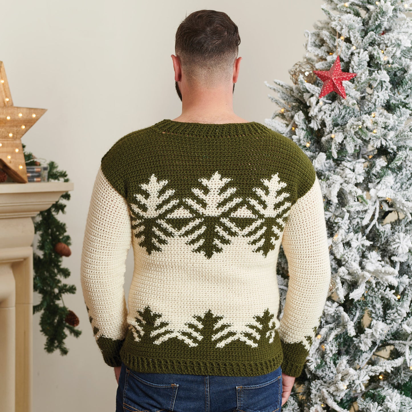 Man in a green and cream holiday sweater with a snowflake pattern stands back to camera. Decorated Christmas tree and lit star are visible. Cozy, festive vibe.
