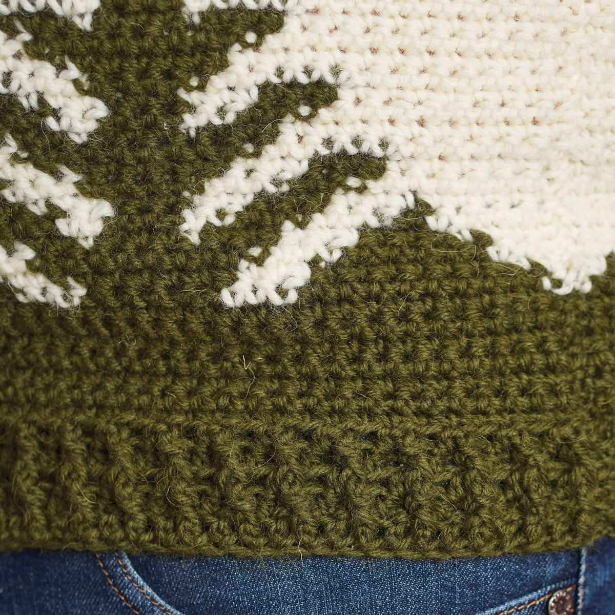 Close-up of a knitted green and cream sweater with a textured pattern resembling a tree. The edge rests above blue denim jeans, conveying warmth.