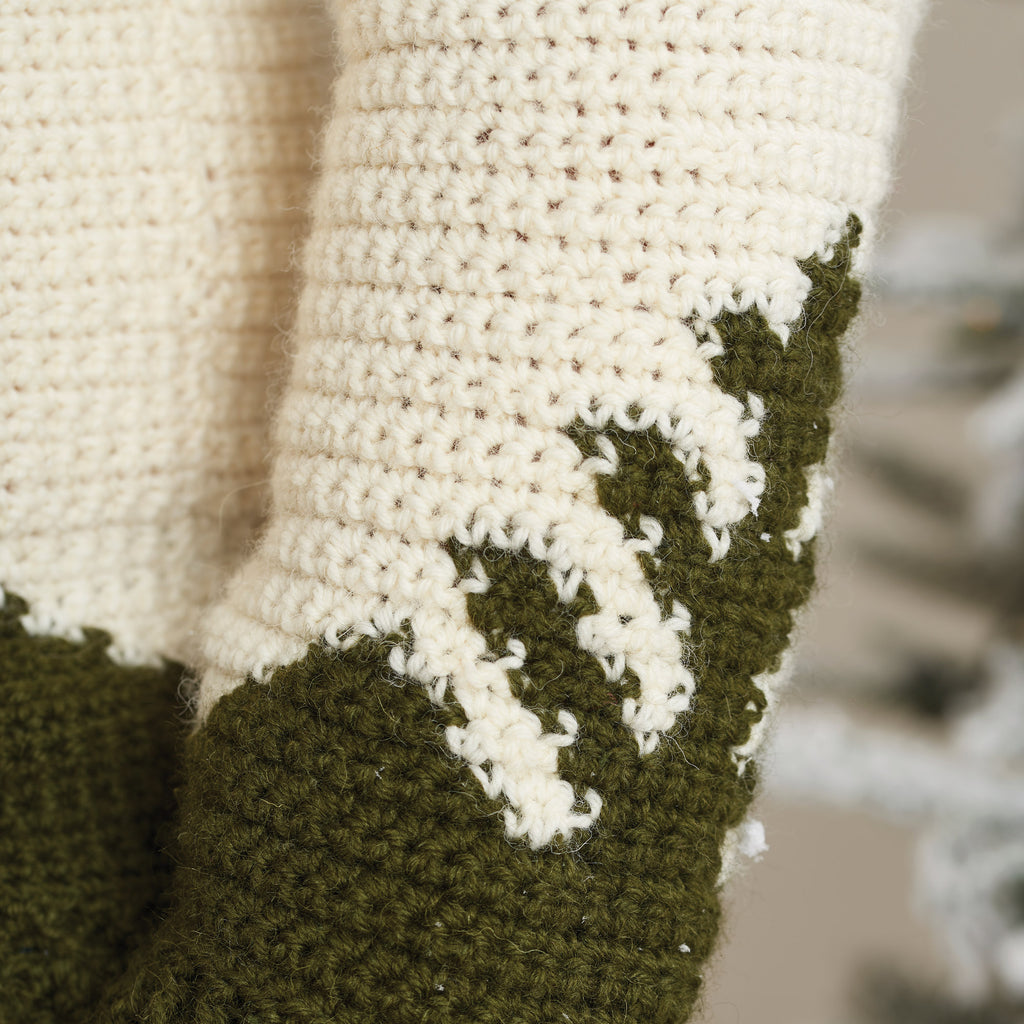 Close-up of a crocheted fabric with cream and green yarn. The pattern resembles mountain peaks, conveying warmth and coziness, set against a blurred background.