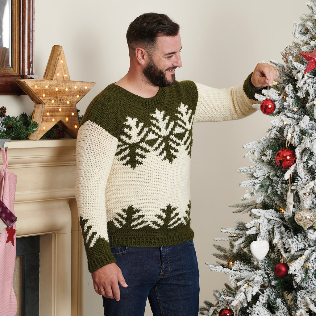 Man smiling while hanging a red ornament on a snowy Christmas tree. He wears a green and white sweater with a festive design. Cozy, holiday atmosphere.