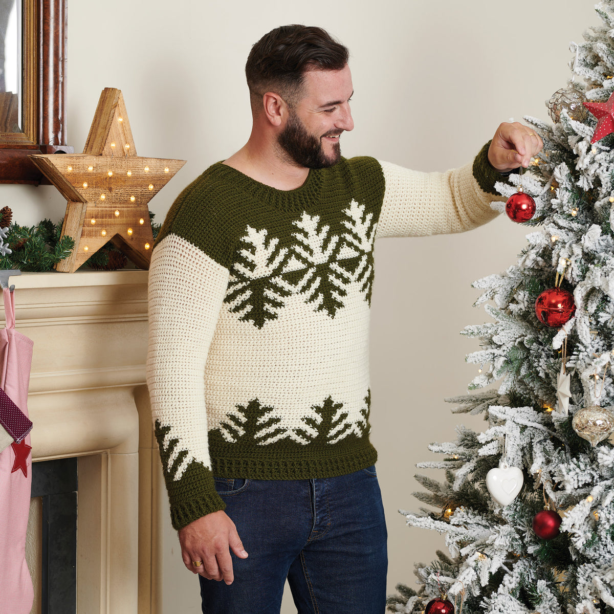 Man smiling while hanging a red ornament on a snowy Christmas tree. He wears a green and white sweater with a festive design. Cozy, holiday atmosphere.