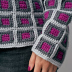 Close-up of a gray and purple crocheted blanket held by a person wearing dark jeans.