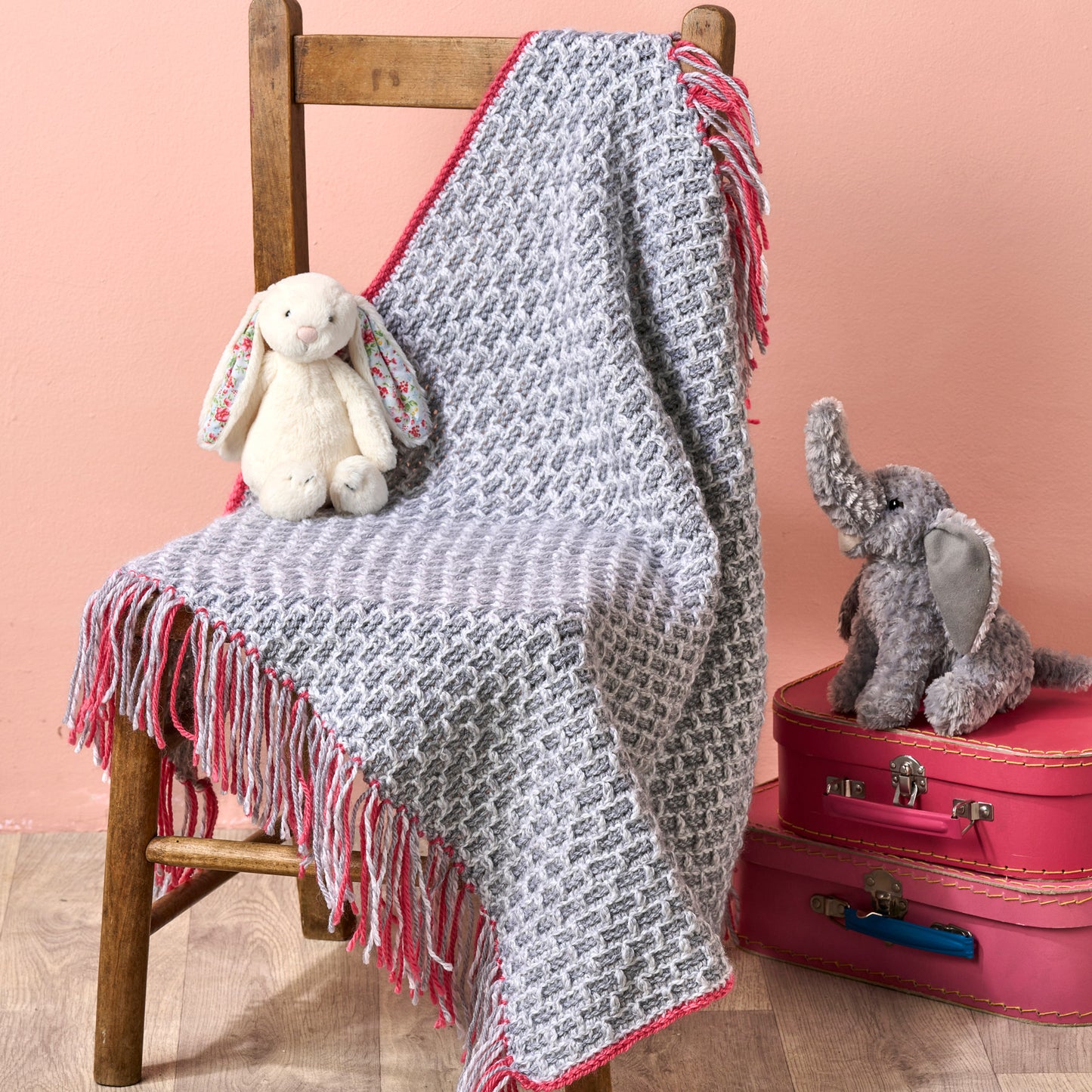 Gray crochet blanket with pink trim draped over a wooden chair, accompanied by stuffed toys on a pink background.