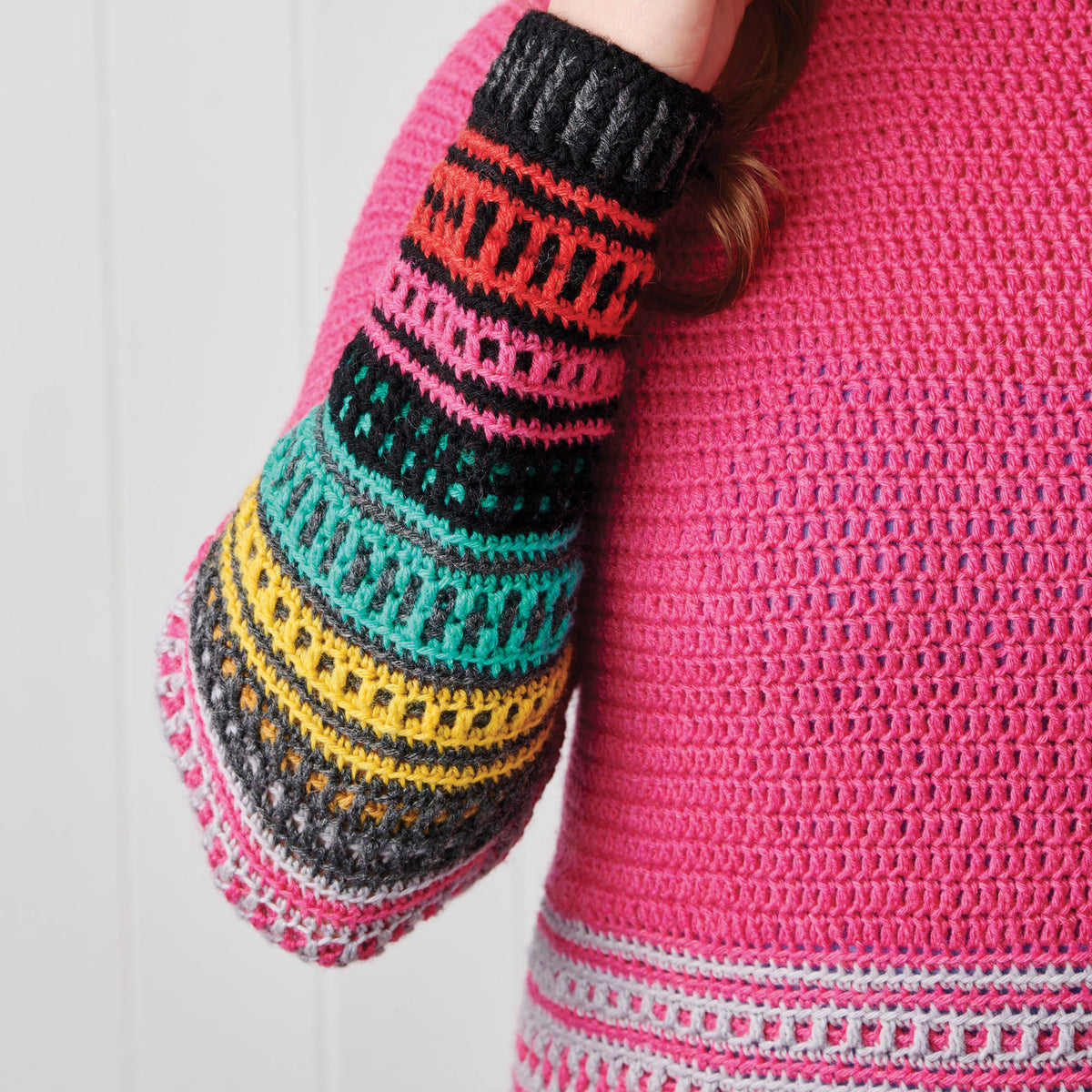 Person wearing a vibrant crochet sweater with a hot pink body and colorful striped sleeve in black, orange, pink, teal, and yellow patterns.
